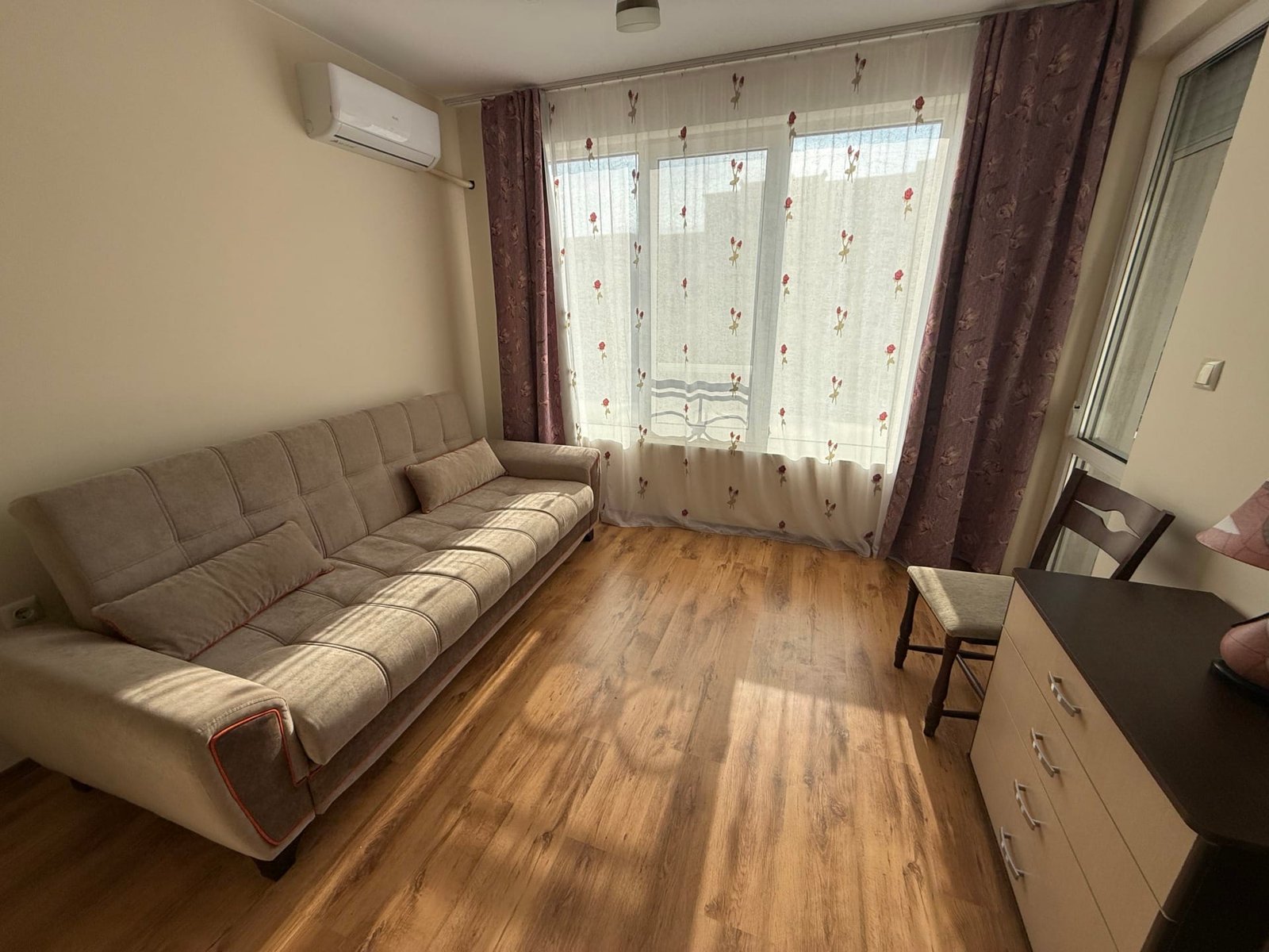 Комфортная гостиная с диваном, деревянным полом и большими окнами, оформлена в светлых тонах для продажи в Болгарии.