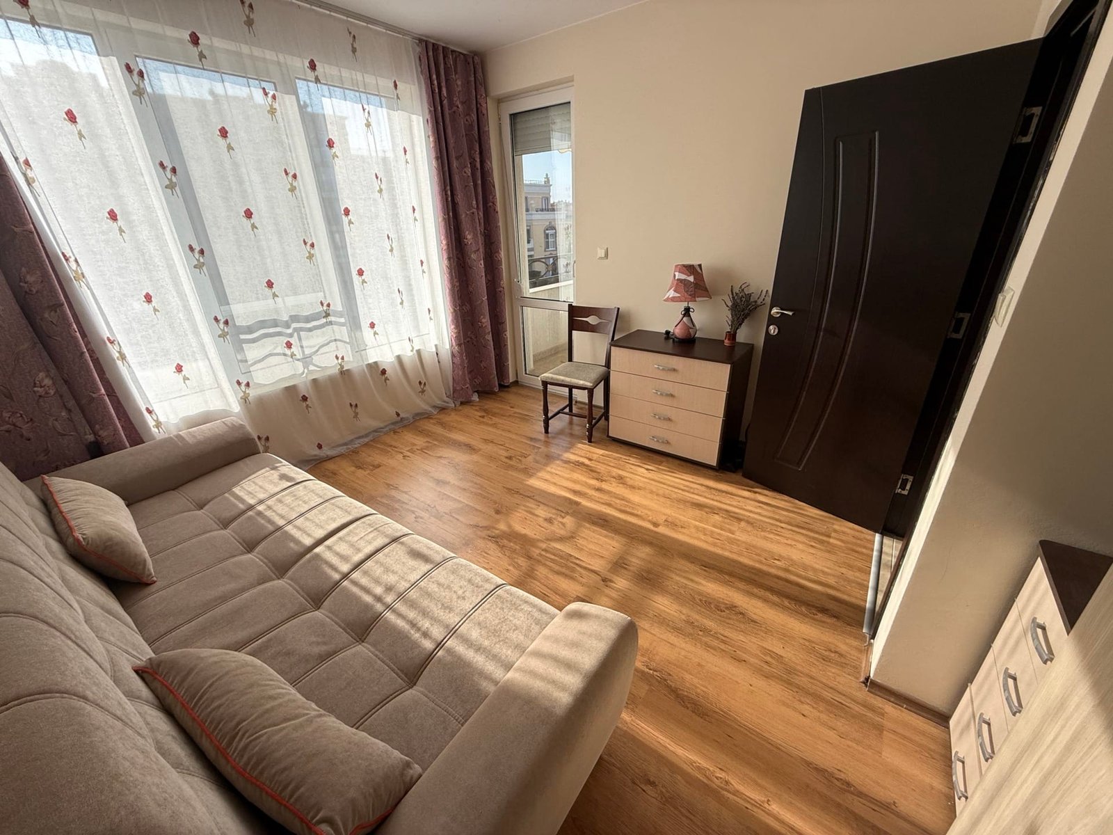 Светлое жилое пространство с секционным диваном, деревянным полом и окнами, выходящими на балкон в Болгарии.