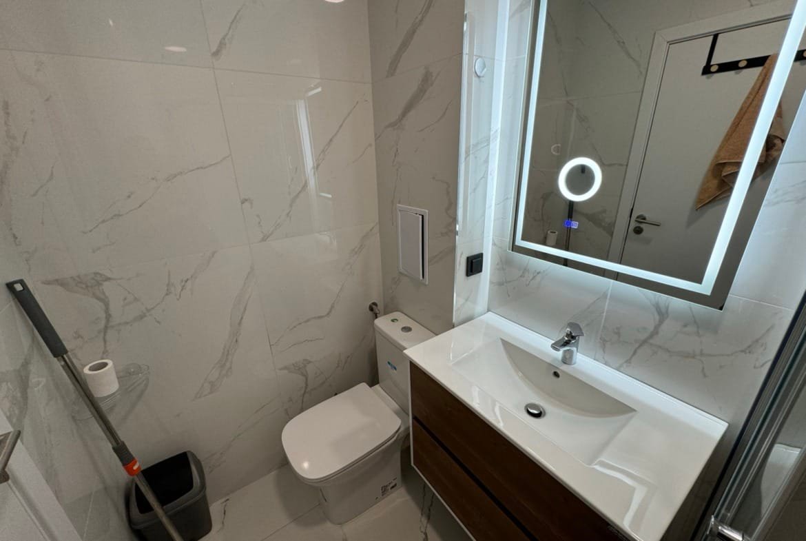 Современный санузел с белой плиткой, стильным зеркалом и сантехникой, подходящий для продажи недвижимости в Болгарии.