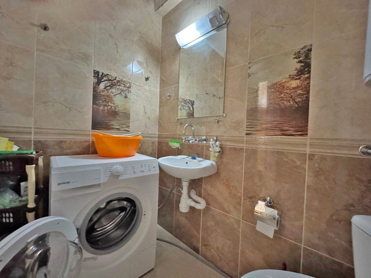 Санузел с ванной машиной, моечной раковиной и зеркалом, отделка под плитку, светлые тона, идеален для квартиры в Болгарии.