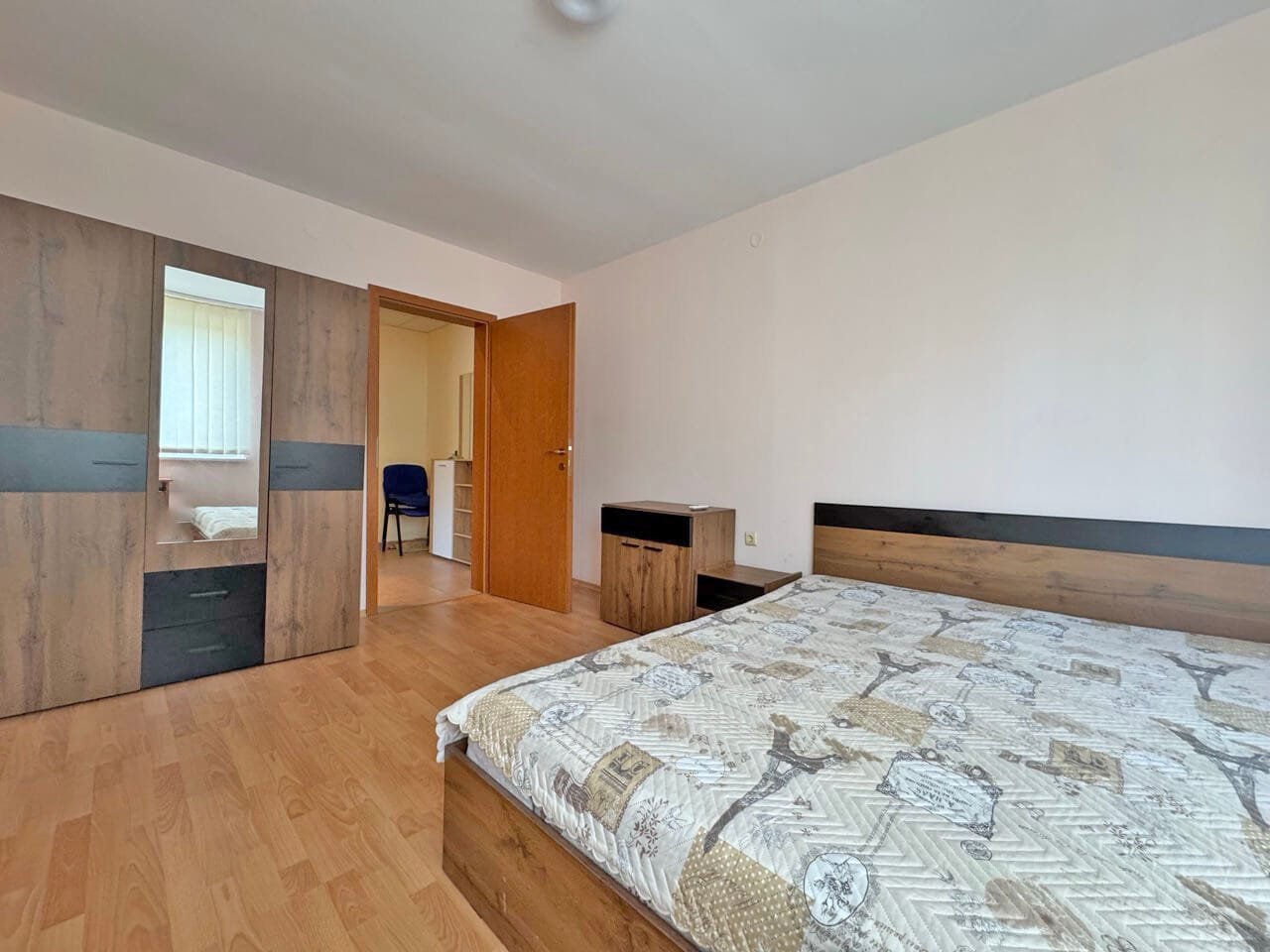 Уютная спальня с двуспальной кроватью и светлым интерьером Спальня с двуспальной кроватью, деревянной мебелью, окнами с занавесками и входом в соседнюю комнату.