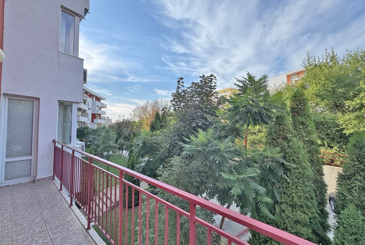 Балкон с красной оградой, зелёные деревья и кустарники, ясное небо, солнечный день в жилом районе.