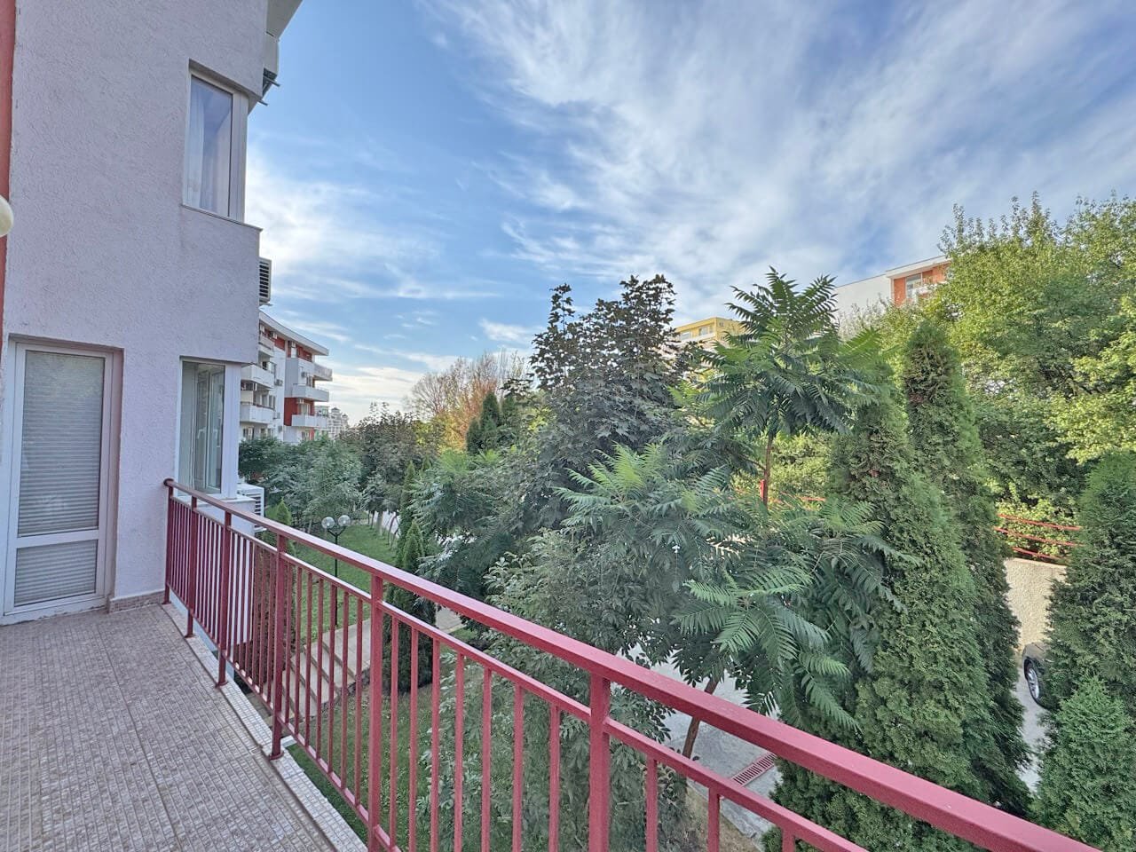 Балкон с зелёным садом и ясным небом в жилом комплексе Балкон с красной оградой, зелёные деревья и кустарники, ясное небо, солнечный день в жилом районе.