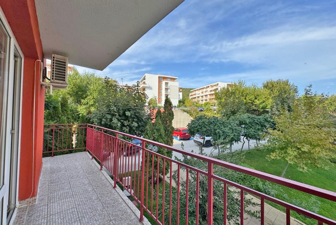 Балкон с красной решеткой, зелеными растениями и видом на жилые здания и парк на фоне ясного голубого неба.