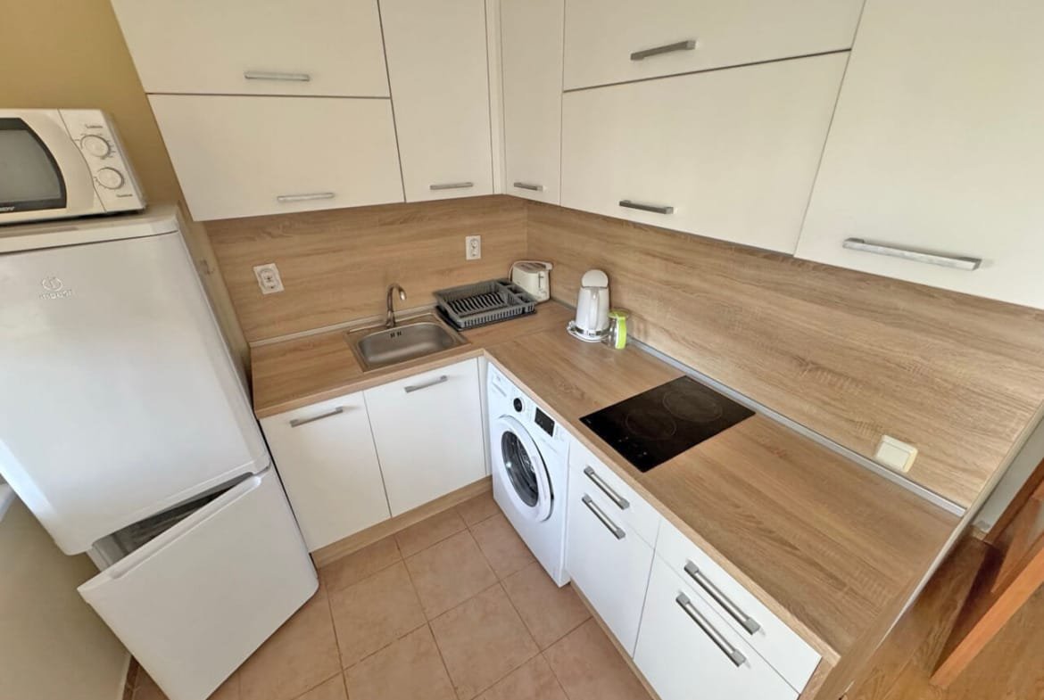 Современная кухня с белыми шкафами, деревянной столешницей, стиральной машиной и техникой, включая микроволновку и мойку.