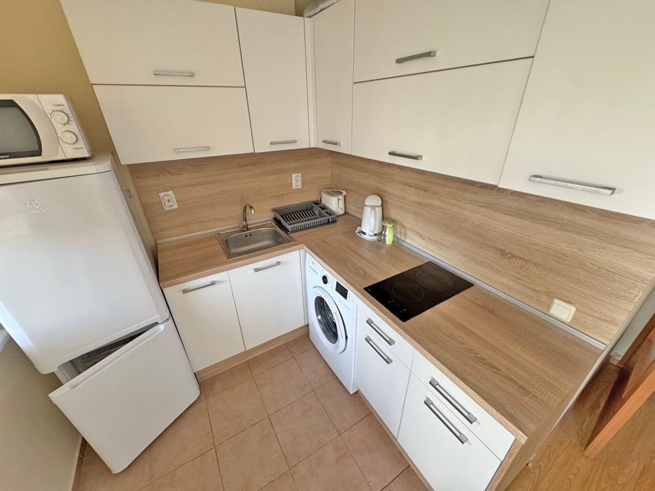 Современная кухня с белыми шкафчиками и бытовой техникой Современная кухня с белыми шкафами, деревянной столешницей, стиральной машиной и техникой, включая микроволновку и мойку.