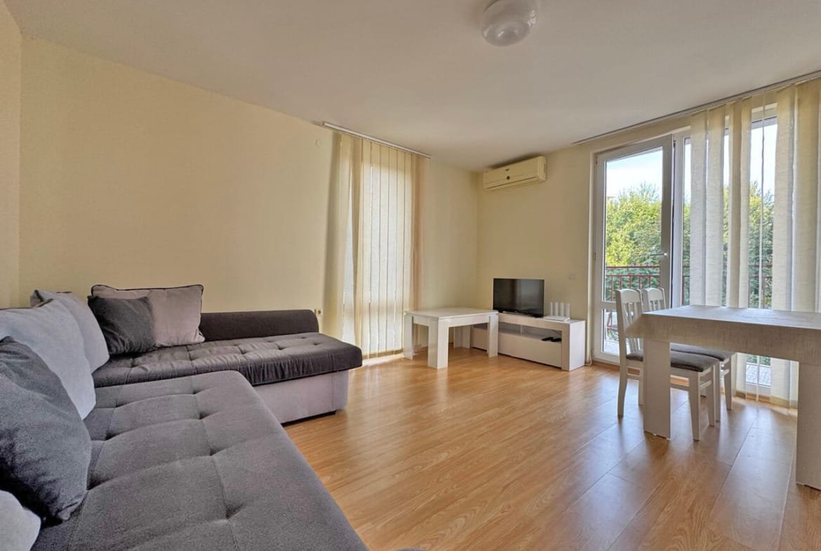 Светлая гостиная с серым диваном, телевизором на белом тумбе, столом и стульями у окна с занавесками.