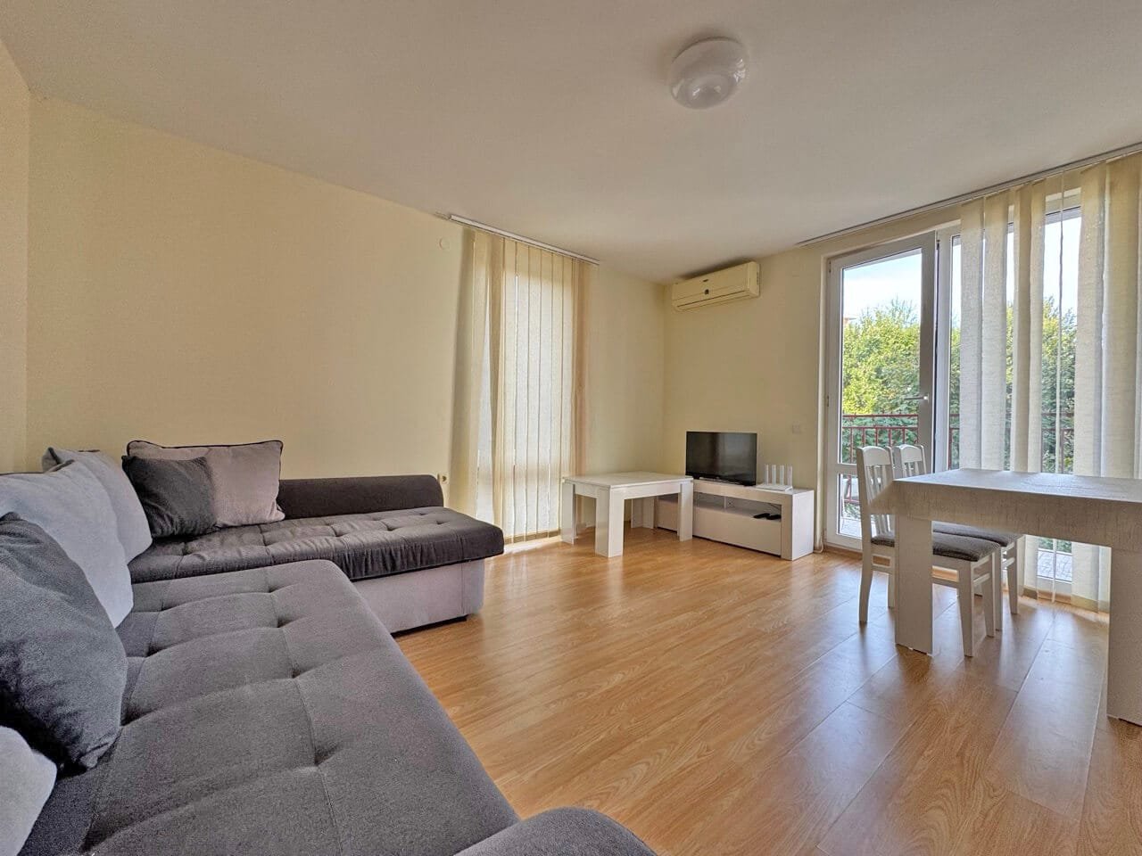 Современная гостиная с диваном и обеденной зоной. Светлая гостиная с серым диваном, телевизором на белом тумбе, столом и стульями у окна с занавесками.