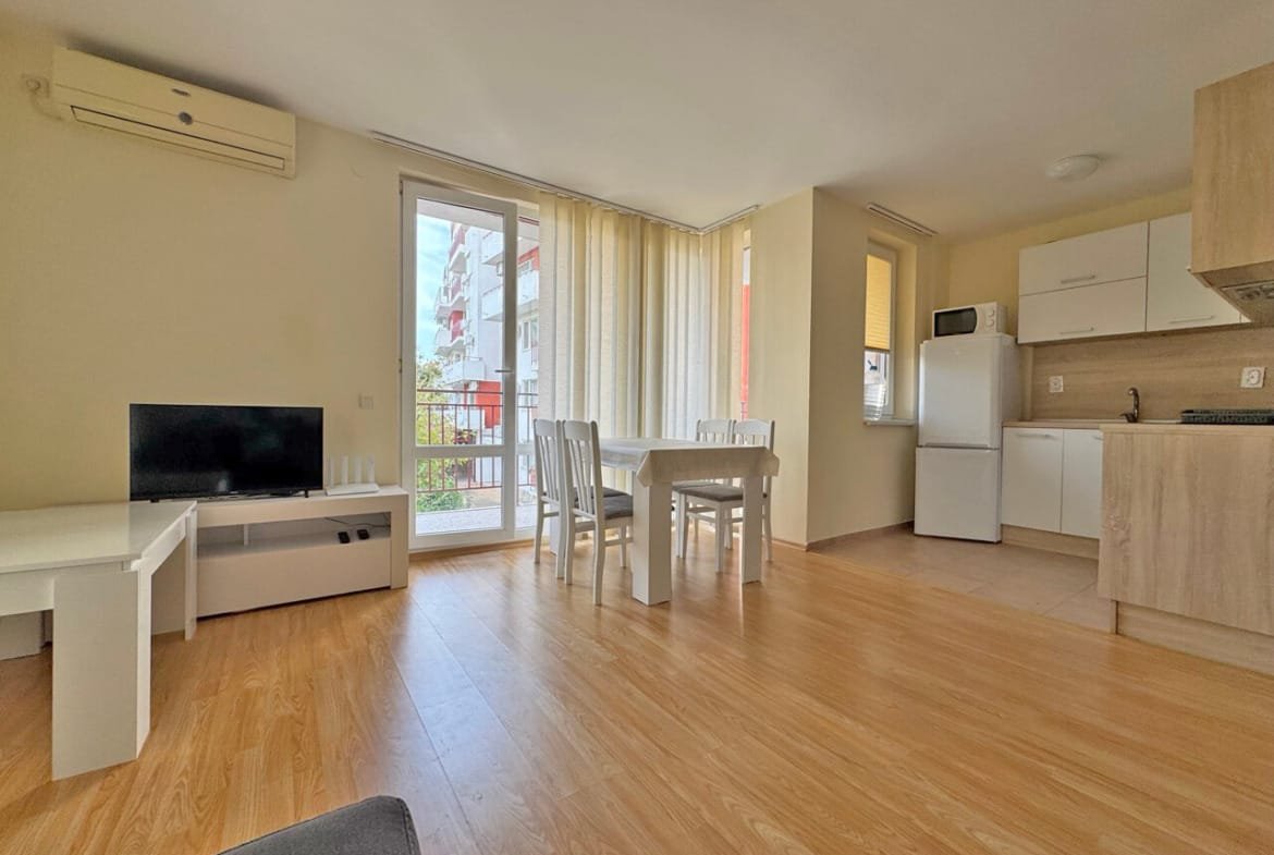 Современная гостиная с обеденным столом, белыми стульями, телевизором и кухней, выходящей на балкон. Светлые тона и деревянный пол.