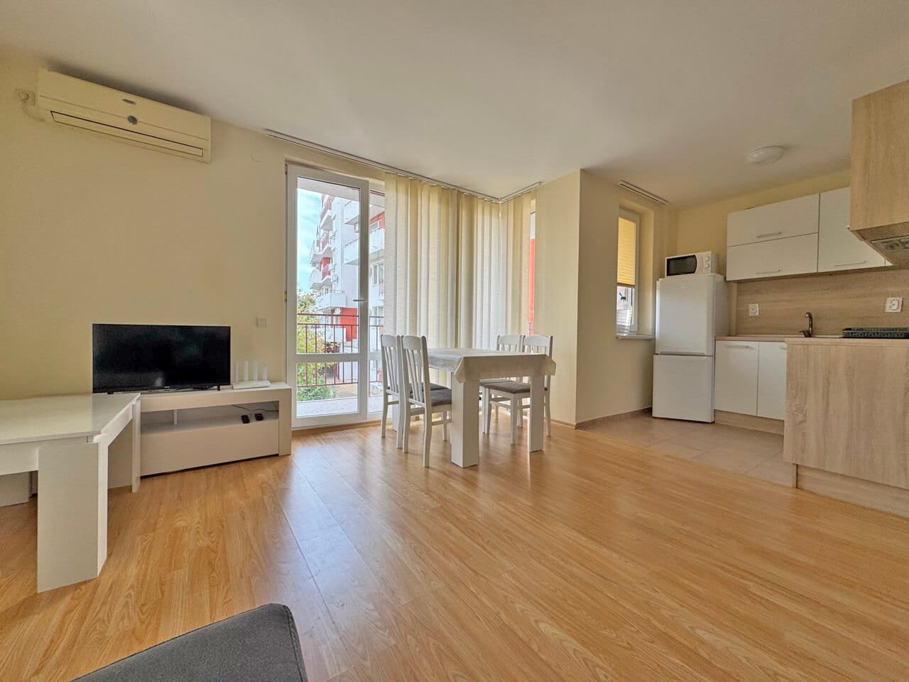 Современный интерьер квартиры с ярким освещением и офисной мебелью Современная гостиная с обеденным столом, белыми стульями, телевизором и кухней, выходящей на балкон. Светлые тона и деревянный пол.