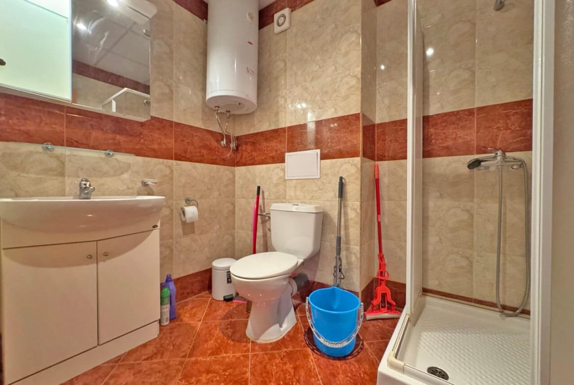 Светлая ванная комната с душевой кабиной, унитазом, раковиной и уборочными принадлежностями. Стены облицованы плиткой.