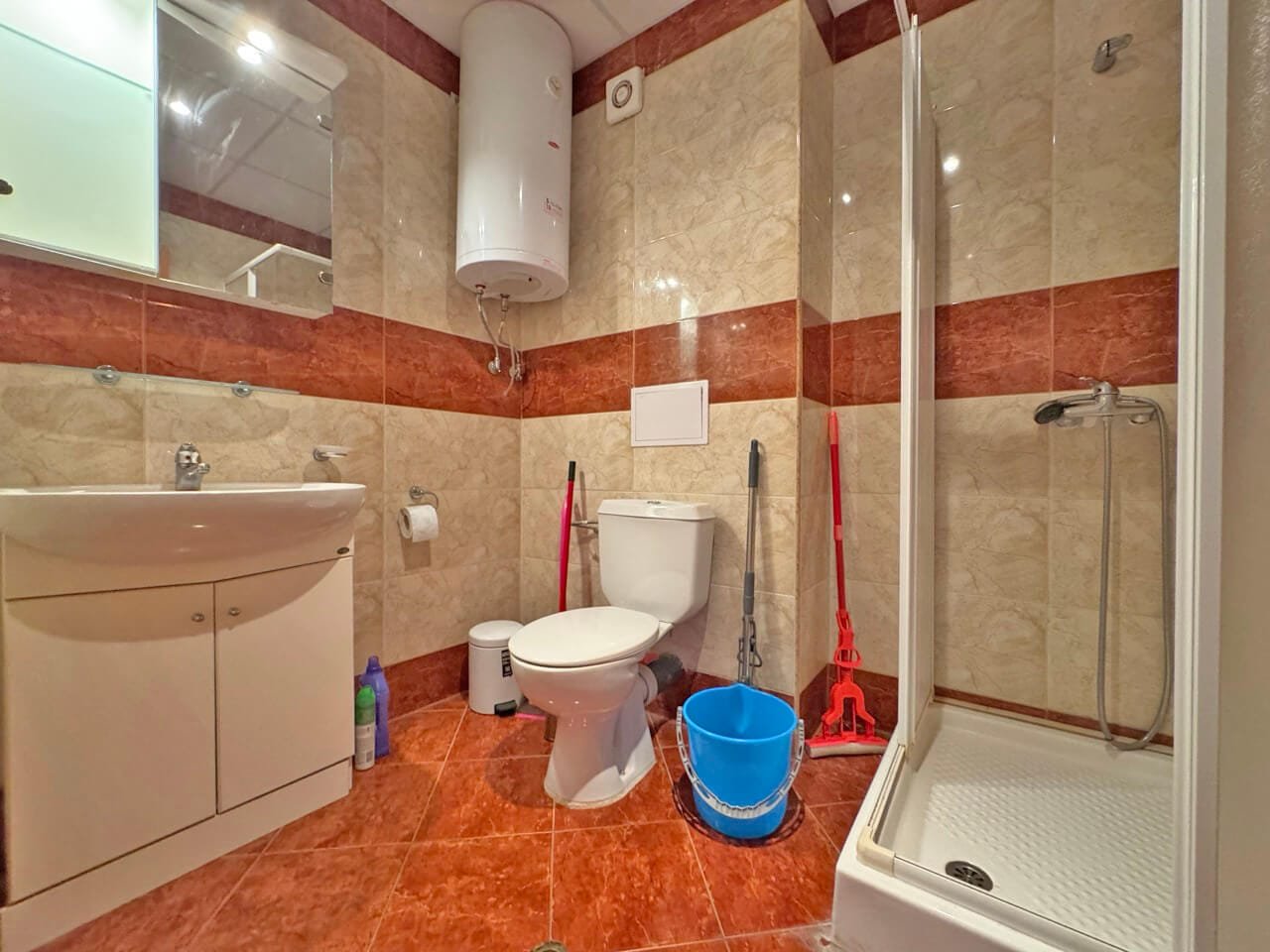 Современная ванная комната с душем и сантехникой Светлая ванная комната с душевой кабиной, унитазом, раковиной и уборочными принадлежностями. Стены облицованы плиткой.