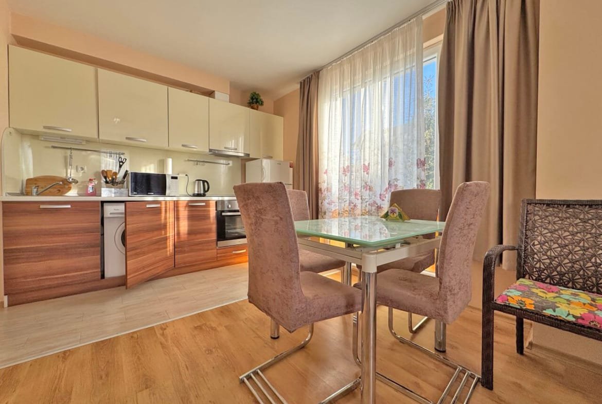 Современная кухня-студия с обеденным столом и стульями, окна с видом на сад, уютная атмосфера для проживания в Болгарии.