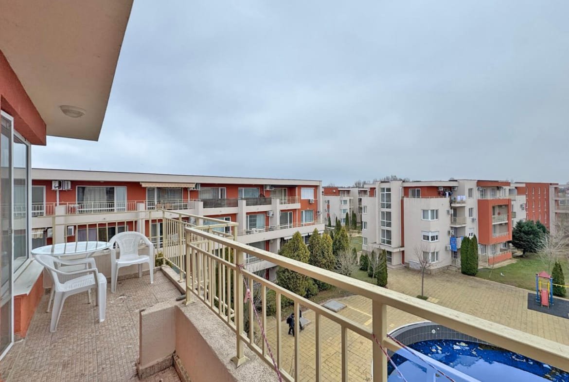 Балкон с белыми пластиковыми столом и стульями, видом на жилые здания и зеленую зону с фонтаном. Погода облачная.