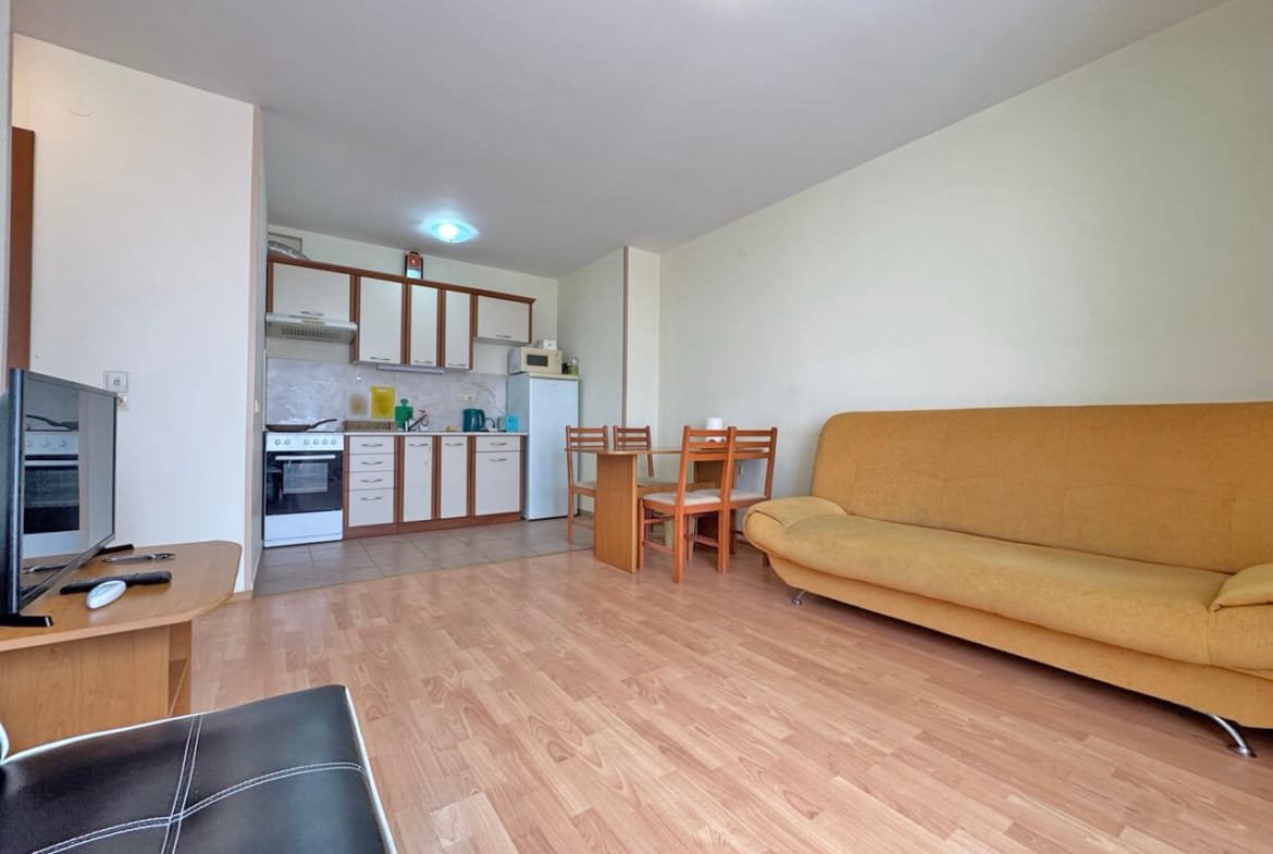 Кухня-студия с обеденной зоной, диваном и техникой, оформленная в светлых тонах и с деревянным полом.