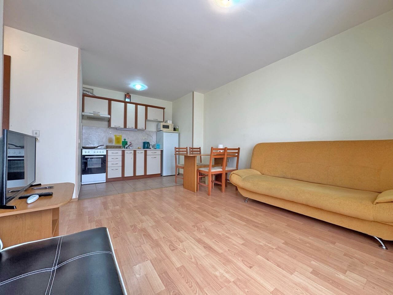 Кухня-студия с обеденной зоной, диваном и техникой, оформленная в светлых тонах и с деревянным полом.