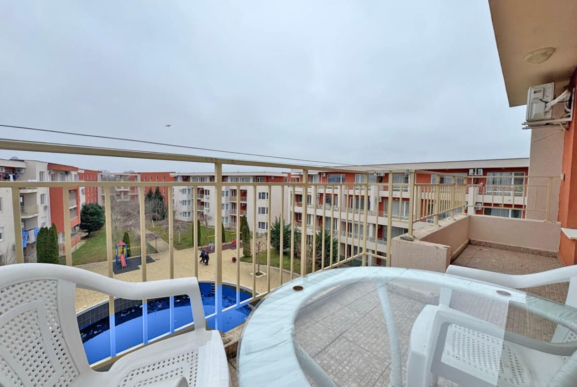 Вид с балкона на жилой комплекс с площадкой для игр, зелеными кустами и облачным небом.