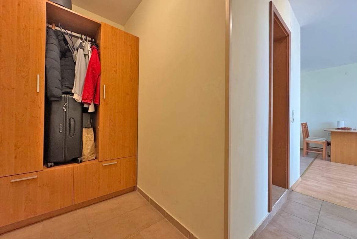 Прихожая с деревянным шкафом, в котором висят куртки и стоят чемоданы, ведущая в светлую комнату с столом.