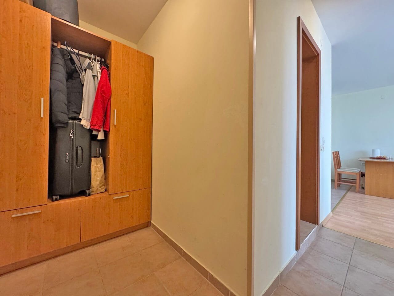 Прихожая с деревянным шкафом, в котором висят куртки и стоят чемоданы, ведущая в светлую комнату с столом.