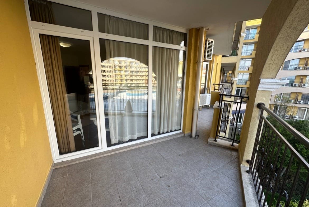 Балкон с плиточным покрытием и стеклянными дверями, вид на соседние здания и зеленую территорию.