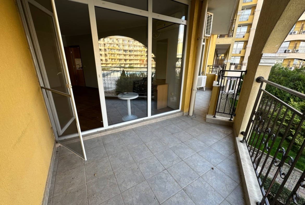 Балкон с плиткой, стеклянной дверью и видом на соседние здания и зелёные насаждения.