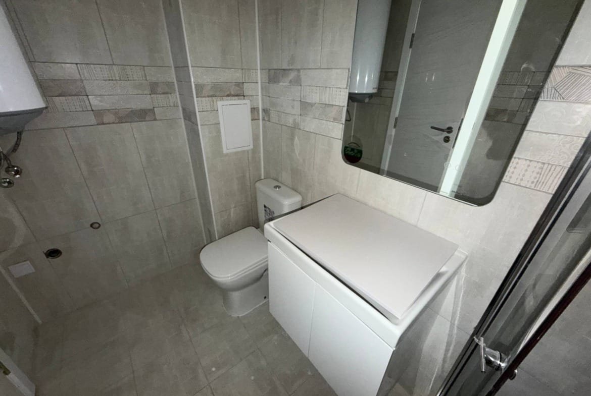 Современный санузел с туалетом, стиральной машиной и зеркалом, отделанный плиткой в светлых тонах.