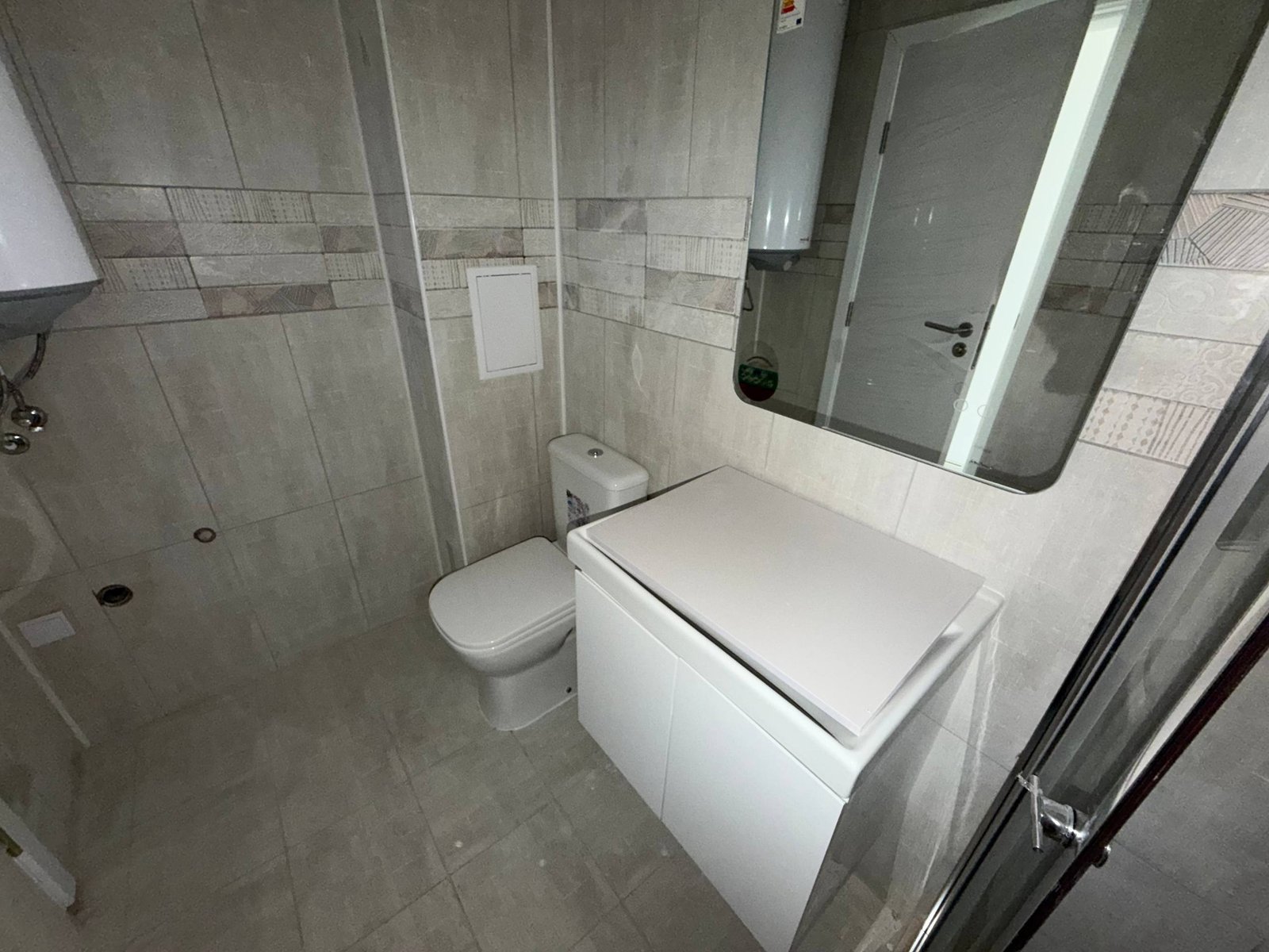 Современный санузел с туалетом, стиральной машиной и зеркалом, отделанный плиткой в светлых тонах.