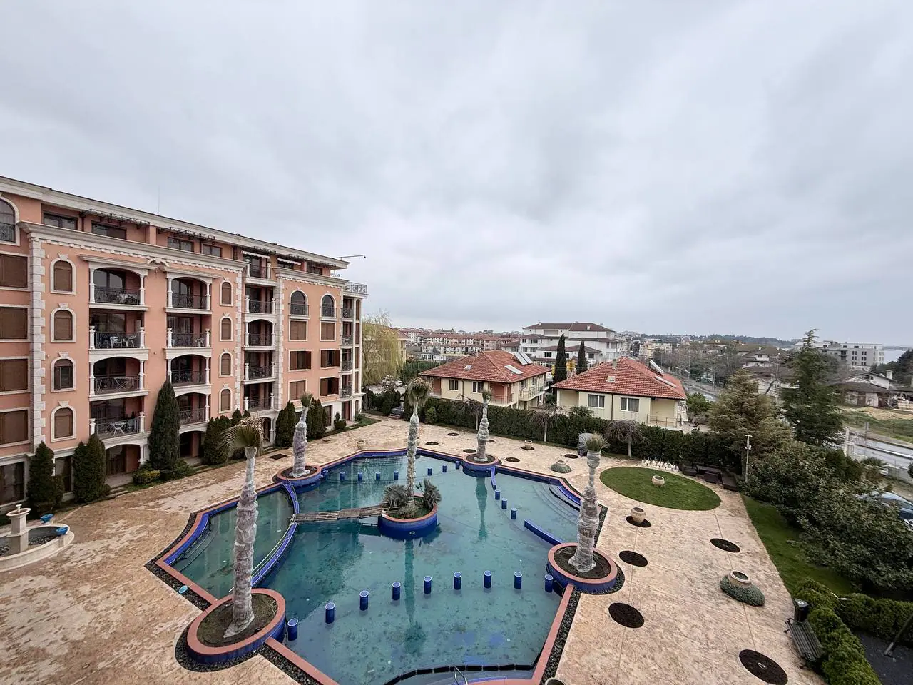 Жилой комплекс с фонтанами и бассейном на фоне облачного неба Жилая недвижимость в Болгарии с бассейном и фонтанами на фоне облачного неба и окружающих вилл.