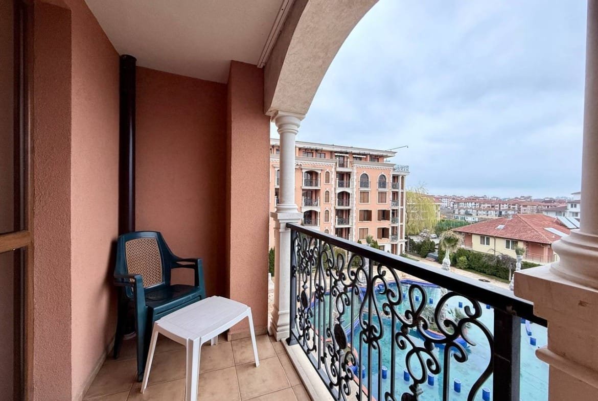 Балкон с пластиковым столом и стулом, вид на жилые комплексы и бассейн в Болгарии, подходящее место для отдыха после покупки недвижимости.