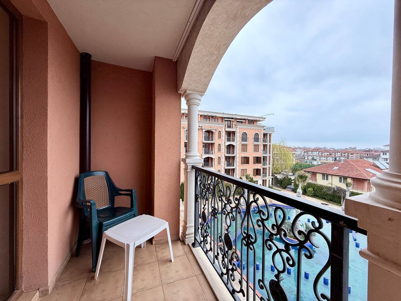 Балкон с видом на бассейн и жилые здания в курортном отеле Балкон с пластиковым столом и стулом, вид на жилые комплексы и бассейн в Болгарии, подходящее место для отдыха после покупки недвижимости.