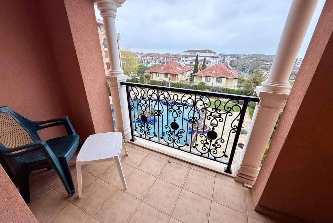 Балкон с черной кованой оградой и пластиковыми стульями, вид на жилые здания и бассейн в Болгарии.