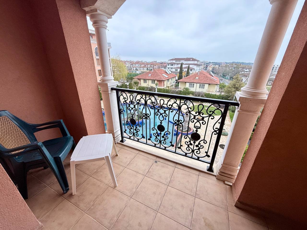 Балкон с видом на пейзаж и бассейн в жилом комплексе. Балкон с черной кованой оградой и пластиковыми стульями, вид на жилые здания и бассейн в Болгарии.