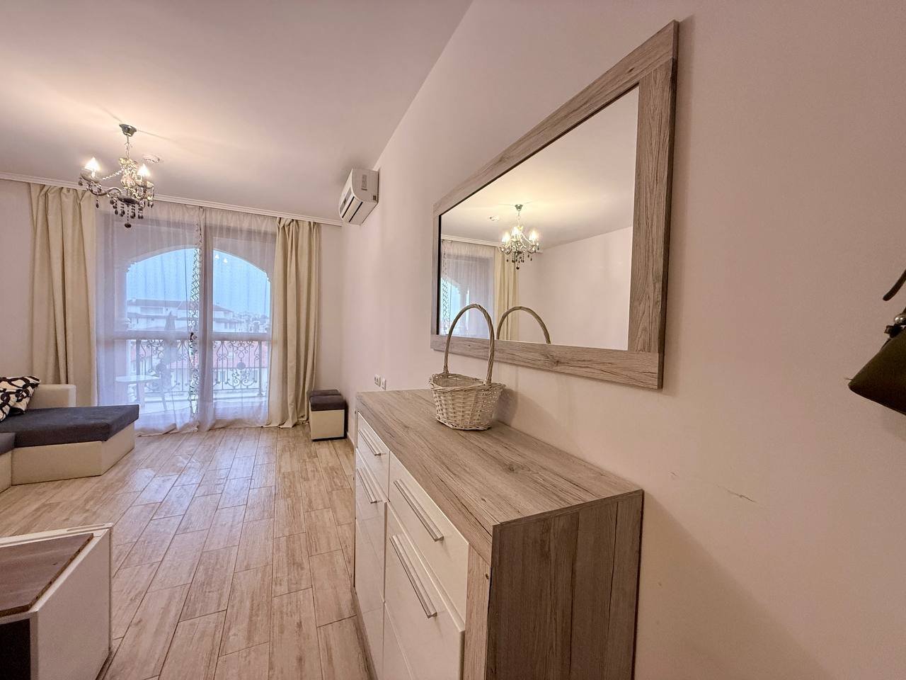 Современный интерьер гостиной с зеркалом и корзиной для хранения Современный интерьер квартиры в Болгарии с белыми стенами, деревянной мебелью и ярким освещением, идеально подходит для жизни или аренды.