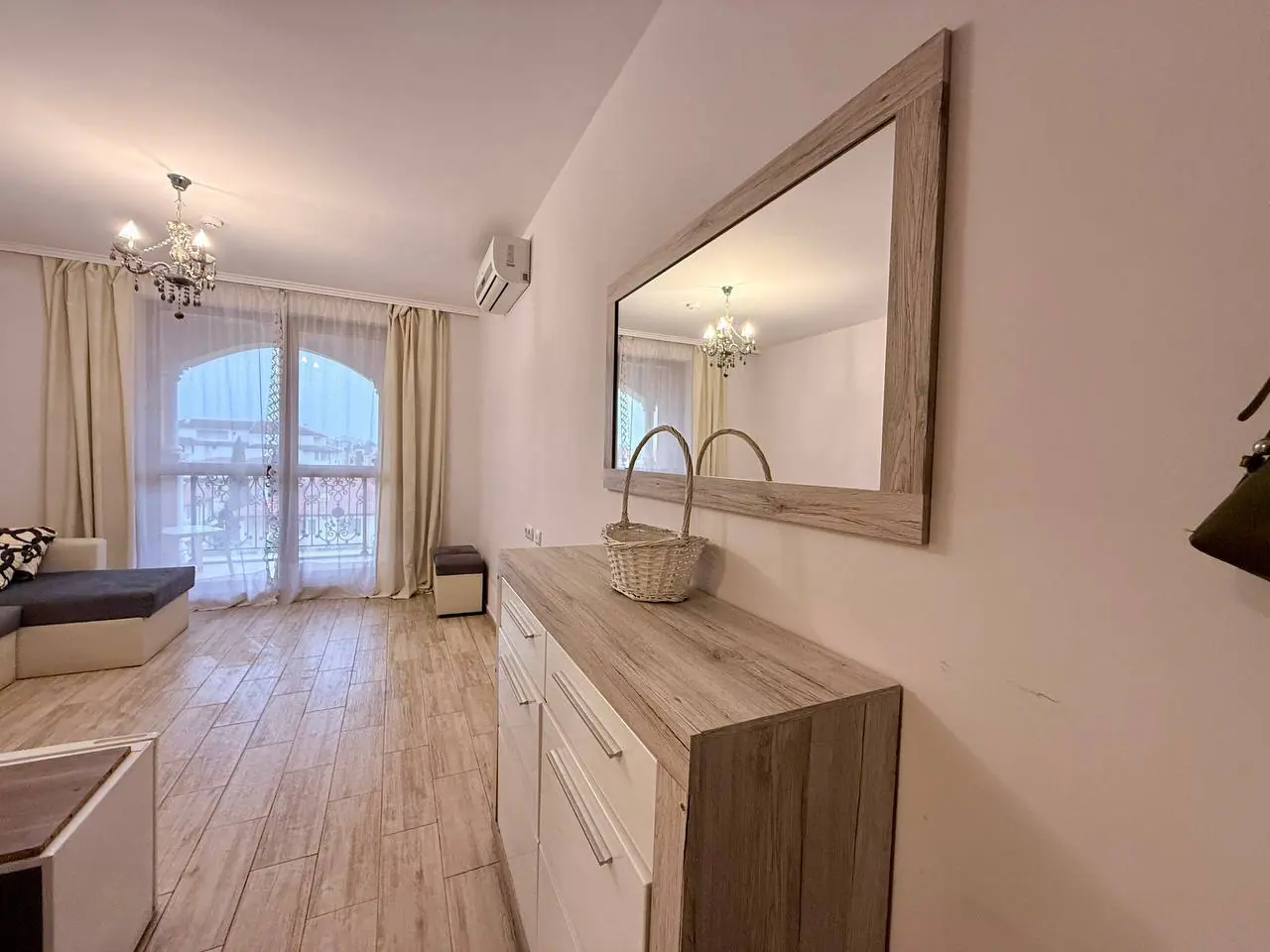 Современный интерьер гостиной с зеркалом и корзиной для хранения Современный интерьер квартиры в Болгарии с белыми стенами, деревянной мебелью и ярким освещением, идеально подходит для жизни или аренды.