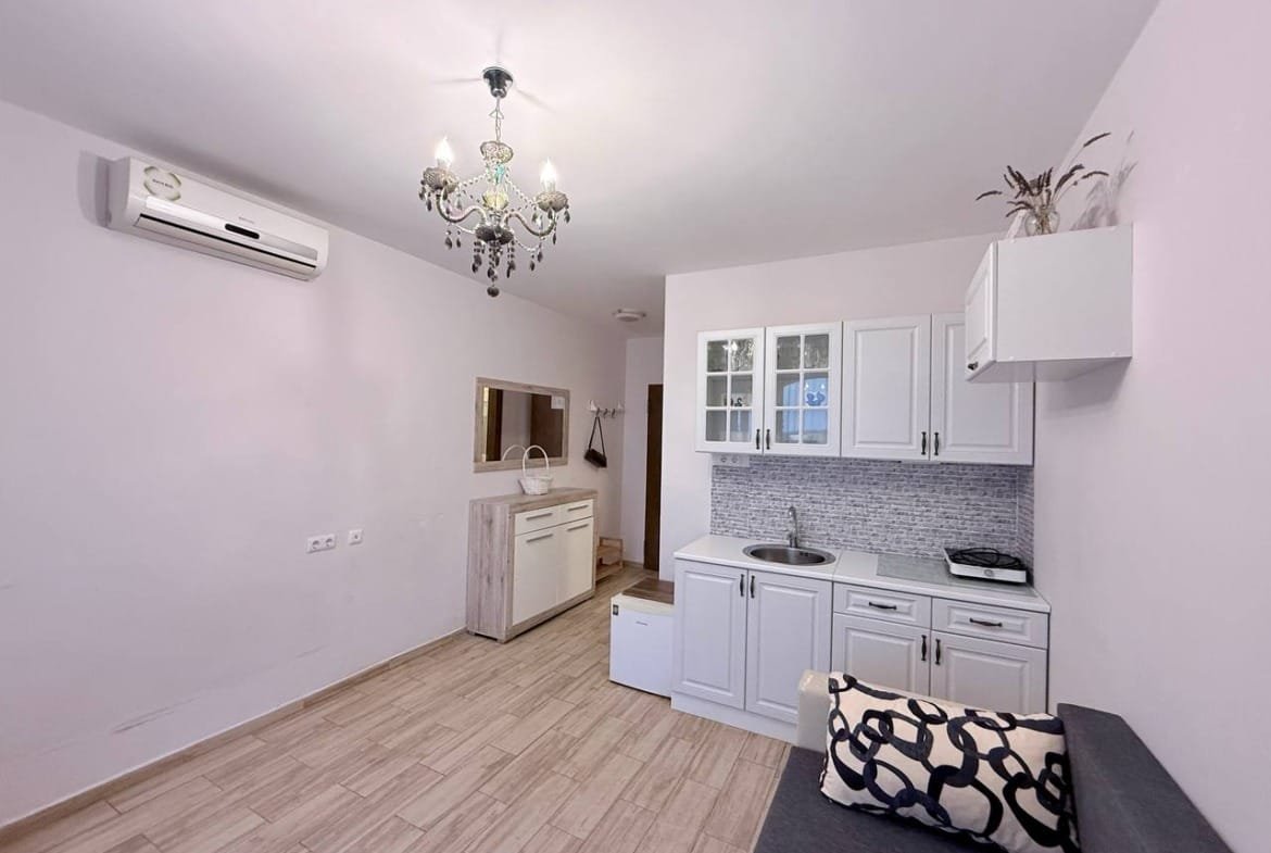 Современная кухня с белыми шкафами и хромированным вентилятором, зона для отдыха с диваном и стильной люстрой в интерьере жилья в Болгарии.