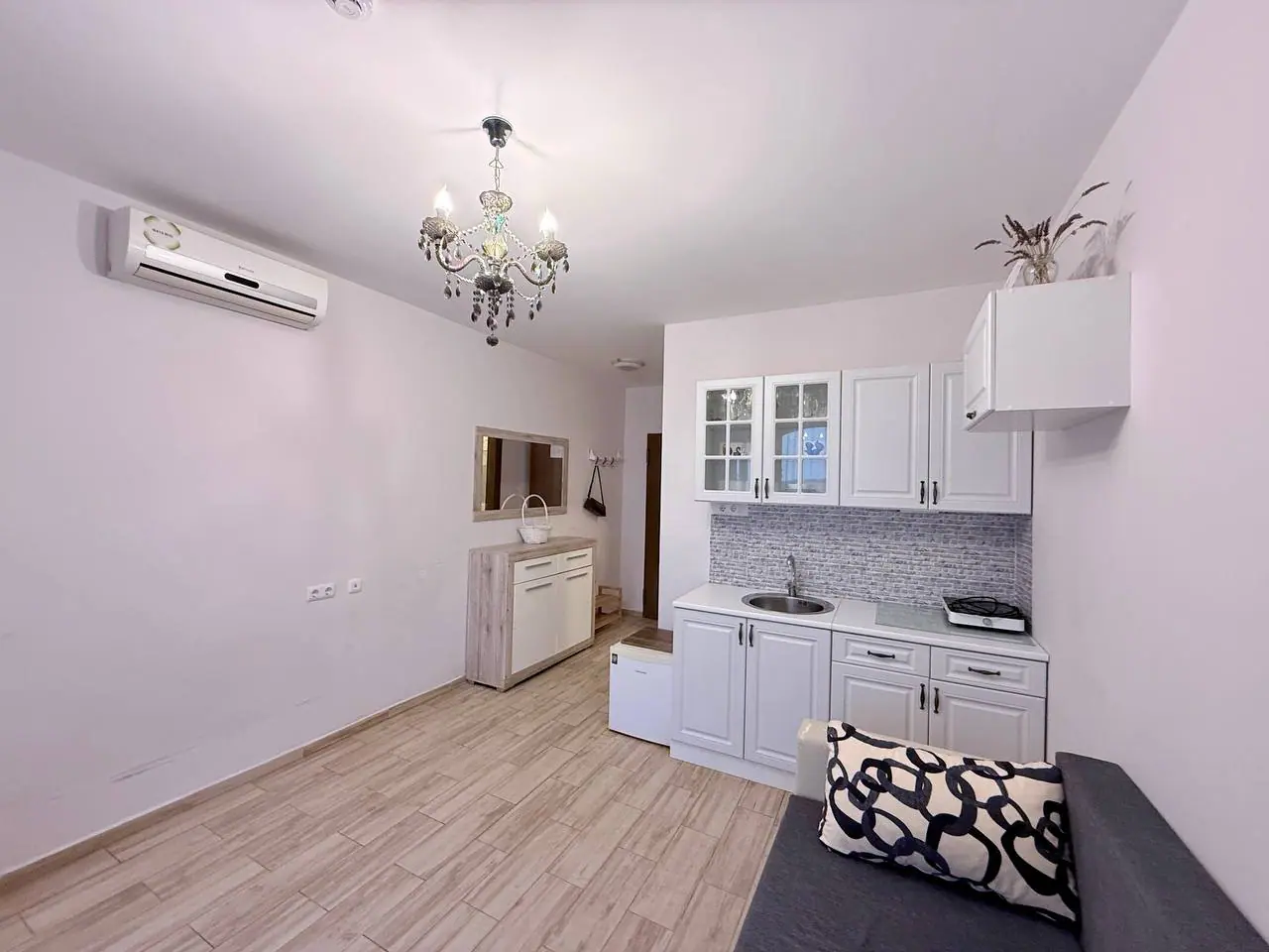 Современная кухня-студия с белыми шкафами и стильным дизайном Современная кухня с белыми шкафами и хромированным вентилятором, зона для отдыха с диваном и стильной люстрой в интерьере жилья в Болгарии.