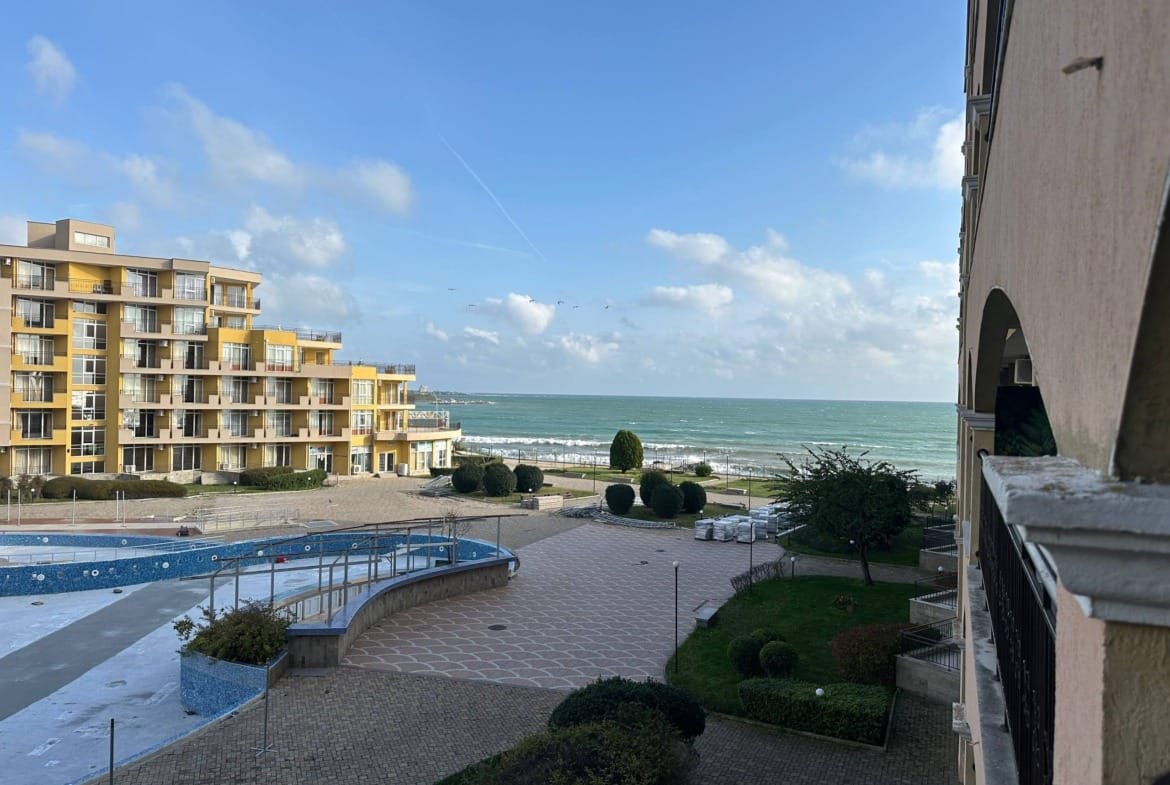 Жилой комплекс у моря с бассейном, зелеными насаждениями и видом на пляж и волну.