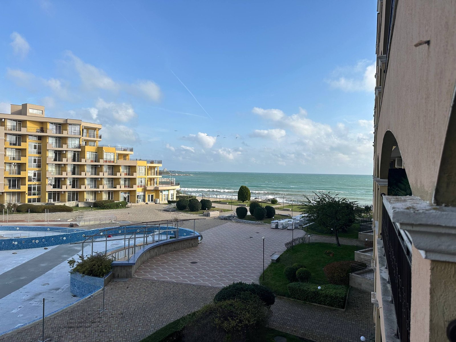 photo_1_2025-12-09_19-05-49 Жилой комплекс у моря с бассейном, зелеными насаждениями и видом на пляж и волну.