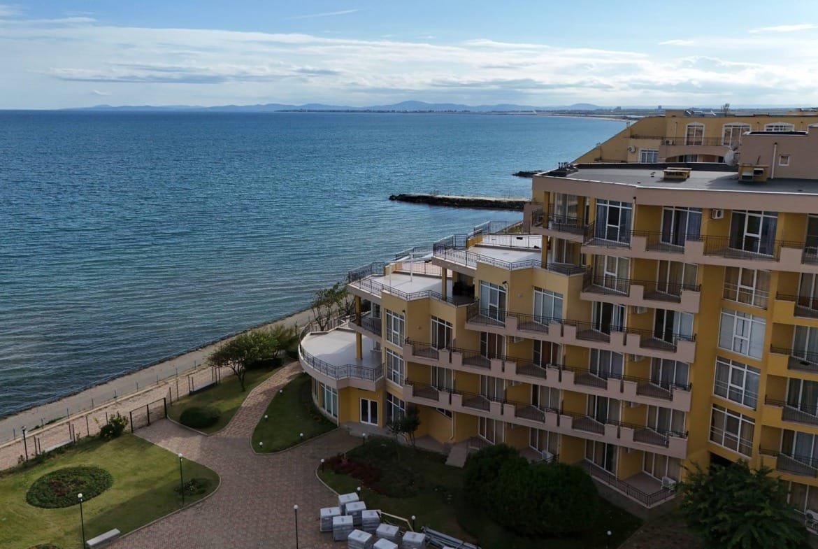 Современный жилой комплекс у моря с зелеными насаждениями и пешеходной дорожкой вдоль пляжа.