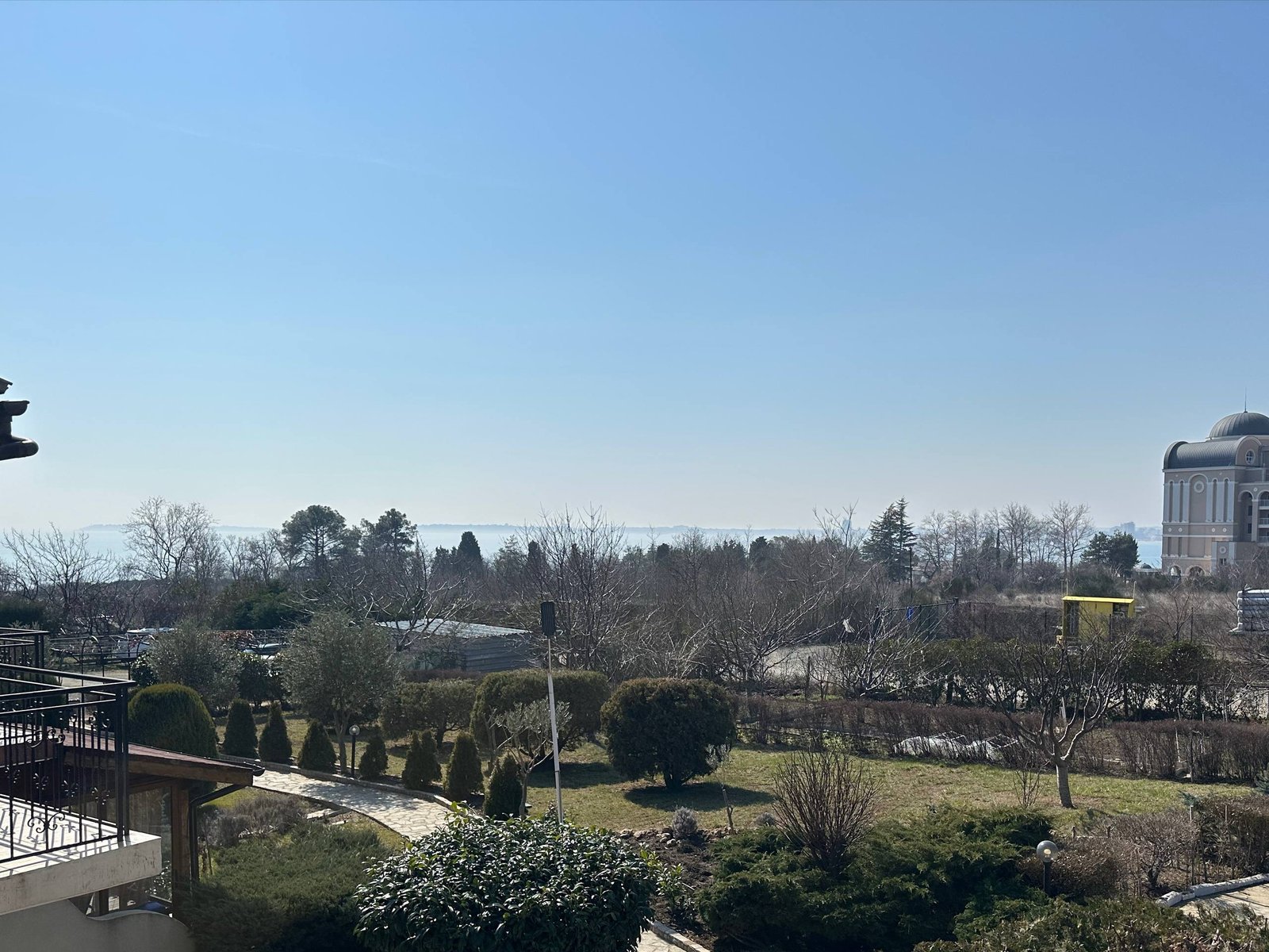 Вид с балкона на зеленый ландшафт, вершины деревьев и море под ясным небом. Создает атмосферу спокойствия и комфорта.