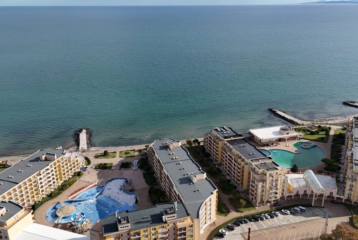 Красивая панорама курортного комплекса с бассейнами и пляжем у моря, спокойная вода и зеленые насаждения.