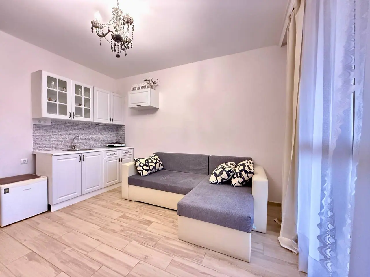 Современная квартира-студия с уютной зоной для отдыха Современная гостиная с серым диваном, кухонным гарнитуром и натуральным освещением, идеальная для комфортного проживания в Болгарии.