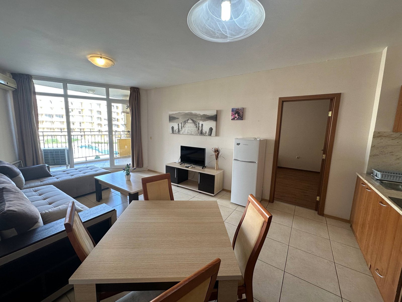 photo_4_2025-12-09_19-05-49 Современная гостиная с кухней, диваном, телевизором и обеденным столом, выход на балкон с видом на улицу.