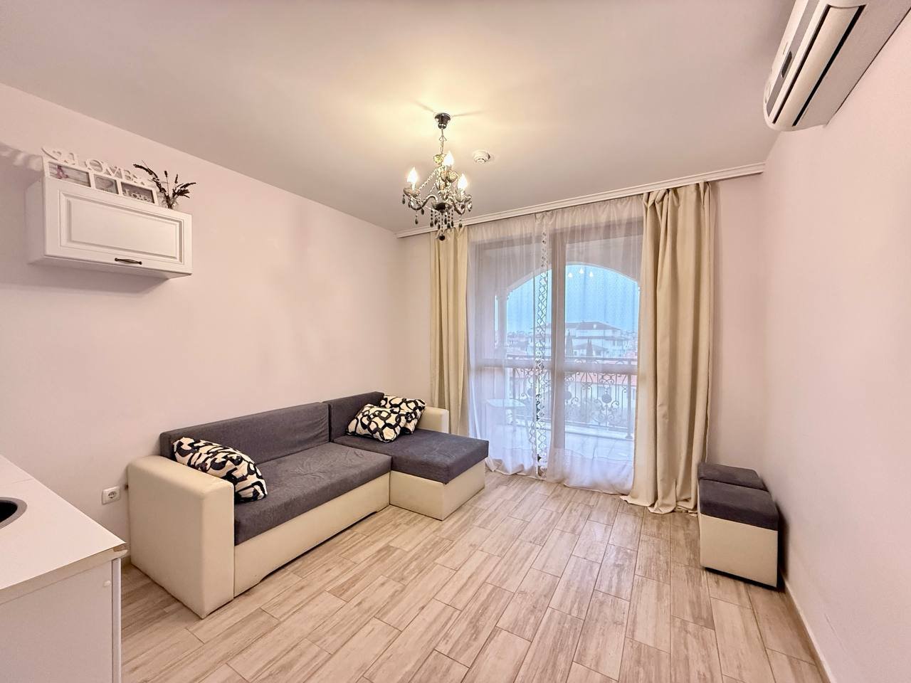 Современная гостиная с диваном и балконом в светлых тонах Современная гостиная с диваном, светлыми стенами и балконной дверью, идеальная для покупки недвижимости в Болгарии.