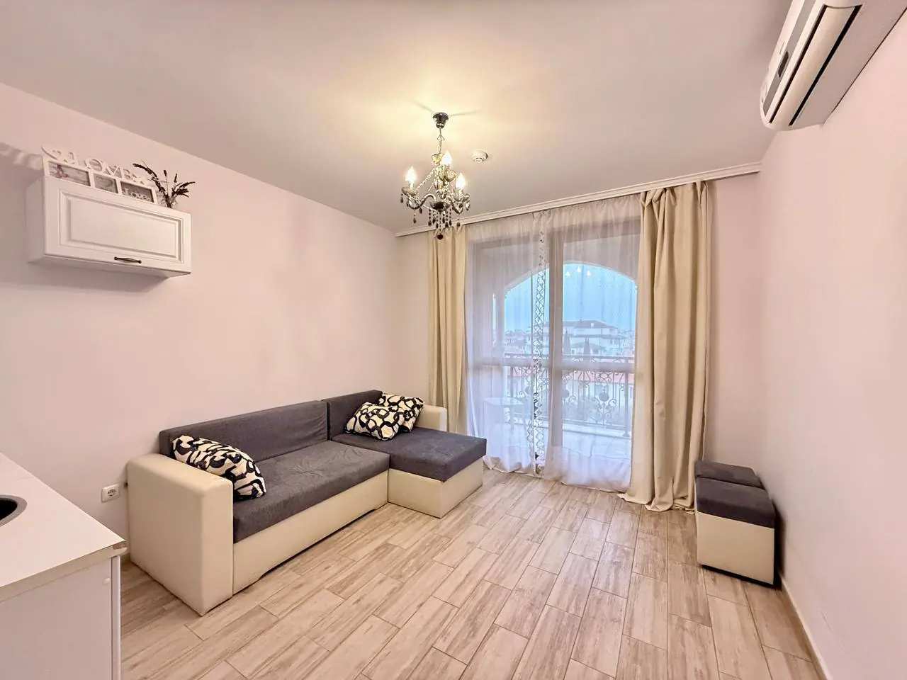 Современная гостиная с диваном и балконом в светлых тонах Современная гостиная с диваном, светлыми стенами и балконной дверью, идеальная для покупки недвижимости в Болгарии.