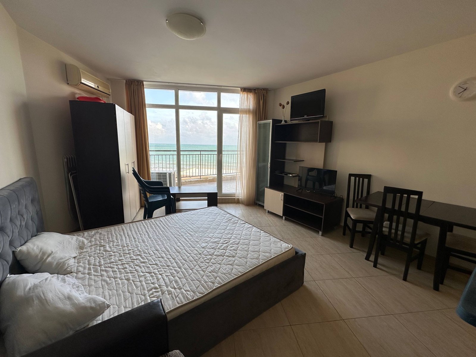 Современная студия с панорамным окном, кроватью, столом, стульями и телевизором, выходящая на море в комплексе “Midia Family Resort”.