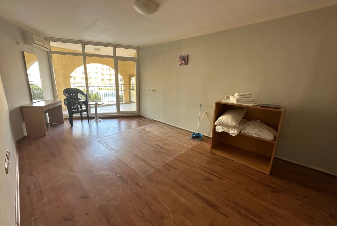 Пустая комната с деревянным полом, столом, стулом и шкафом с постельным бельём, окнами на балкон.