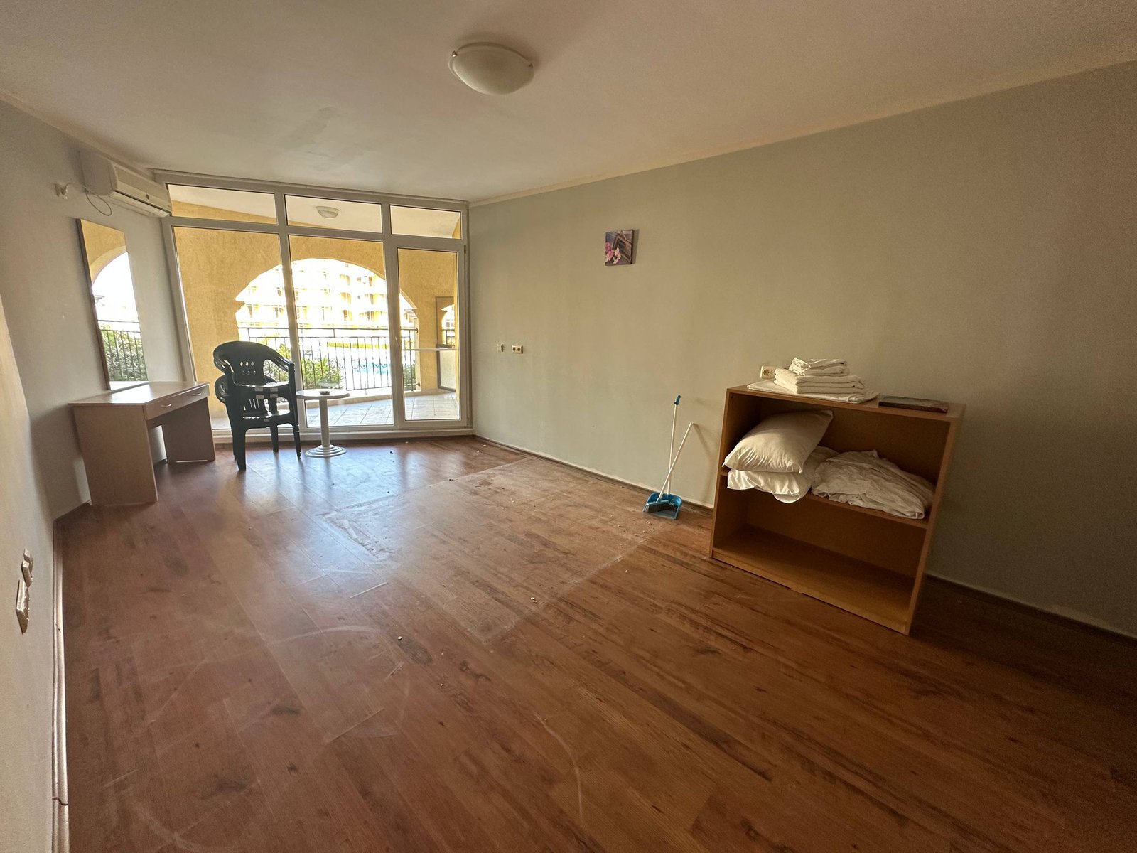 photo_5_2025-12-09_19-10-27 Пустая комната с деревянным полом, столом, стулом и шкафом с постельным бельём, окнами на балкон.