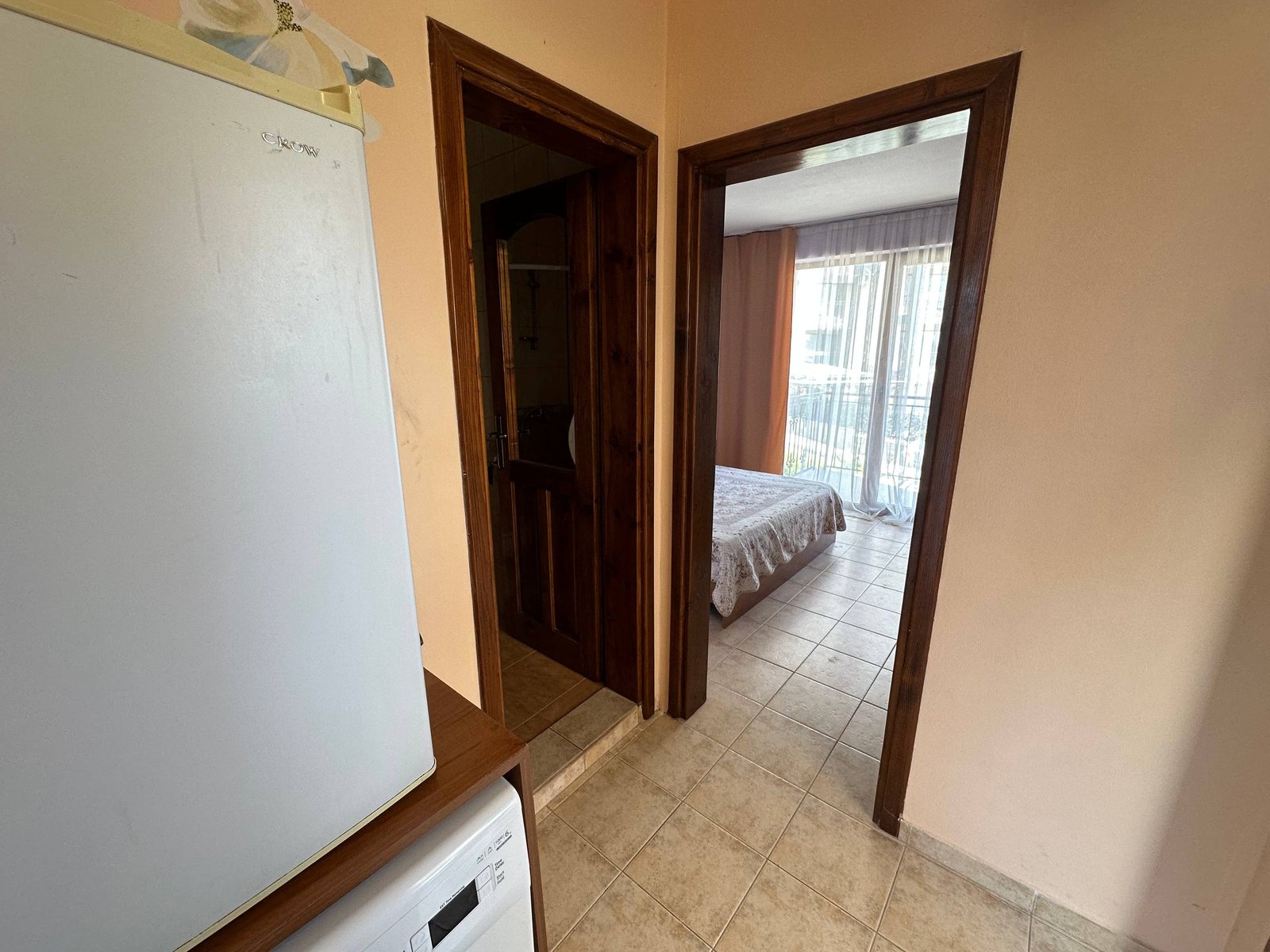 Вход в двухкомнатный апартамент с кухней, ванной и спальней, светлые тона, плиточный пол и большие окна.