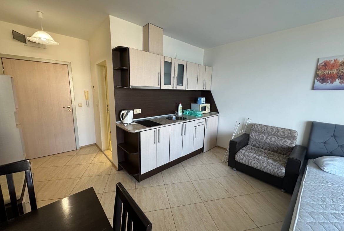 Современная студия с кухней, диваном и обеденным столом; светлые тона, плитка на полу, окно с естественным освещением.